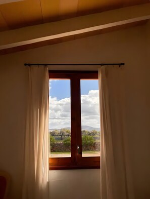 Een kamer met een groot raam, gordijnen en uitzicht op een landschap met bomen en heuvels.