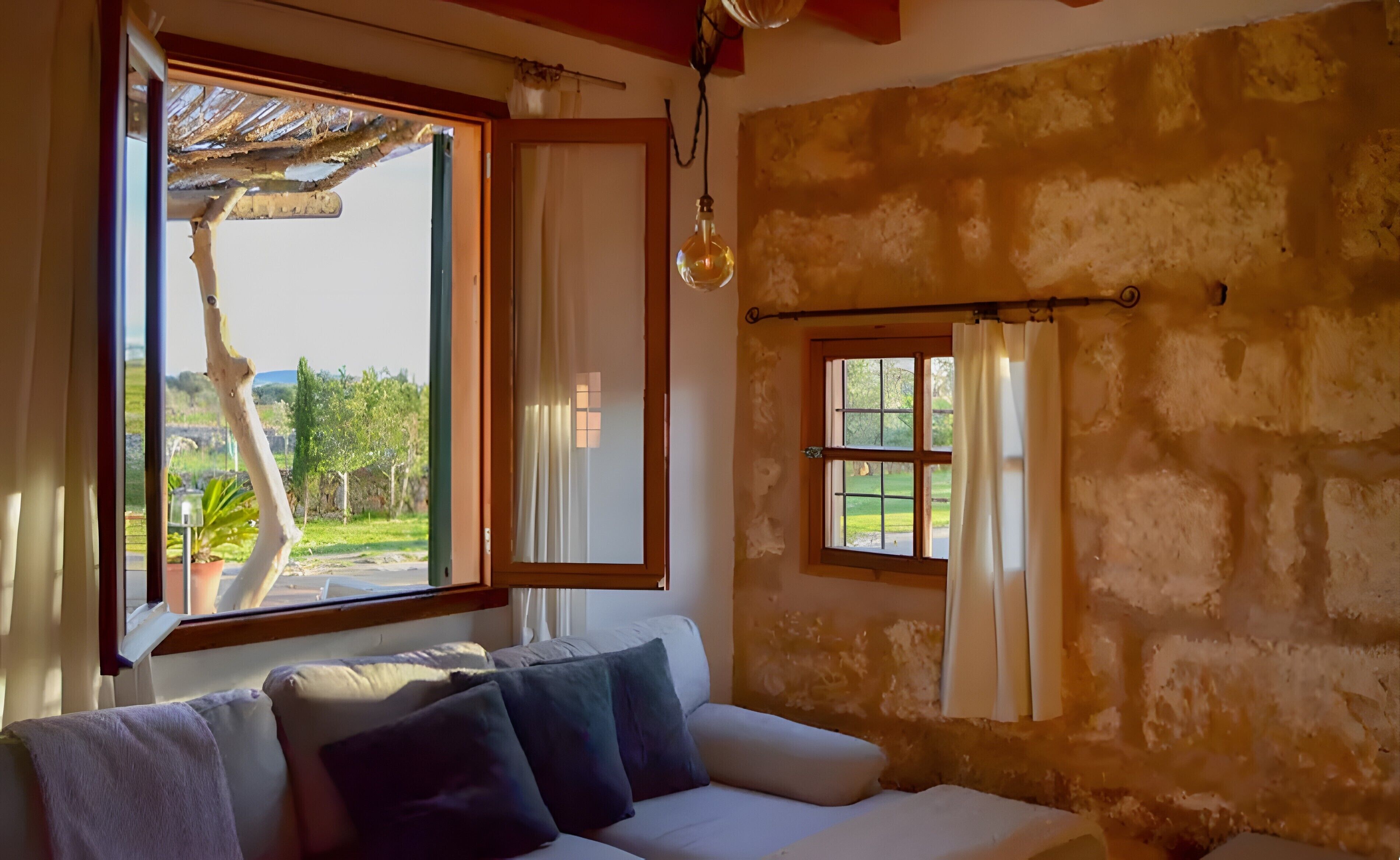 Een kamer met een bank, een raam met gordijnen, een hanglamp en uitzicht op groen.
