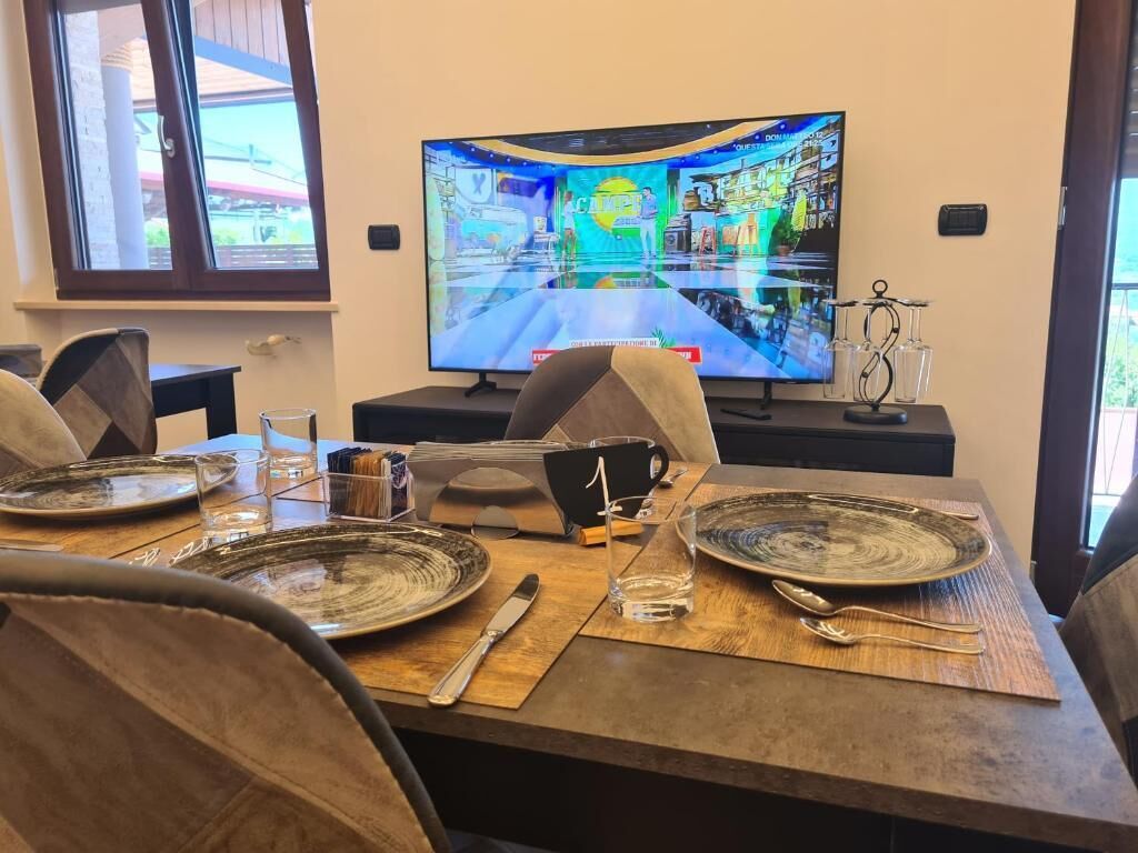 Zona pranzo con tavolo apparecchiato per quattro, televisore che mostra un interno colorato e finestra con vista su vegetazione.