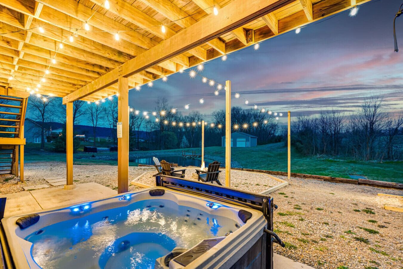 Un bain à remous sous une pergola en bois, illuminé de guirlandes lumineuses, entouré d’une zone recouverte de gravier et offrant une vue sur une maison et des arbres.