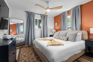 Ein Schlafzimmer mit Bett, Deckenventilator, Nachttisch, Lampe und einem Fenster mit Vorhängen.