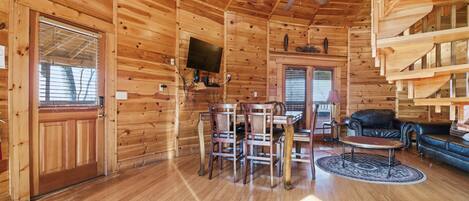 A wooden cabin interior with a dining table, chairs, a sofa, and a ceiling fan.