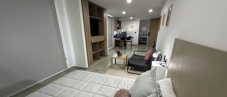 A bedroom with a bed, a desk, and a kitchenette in the background.