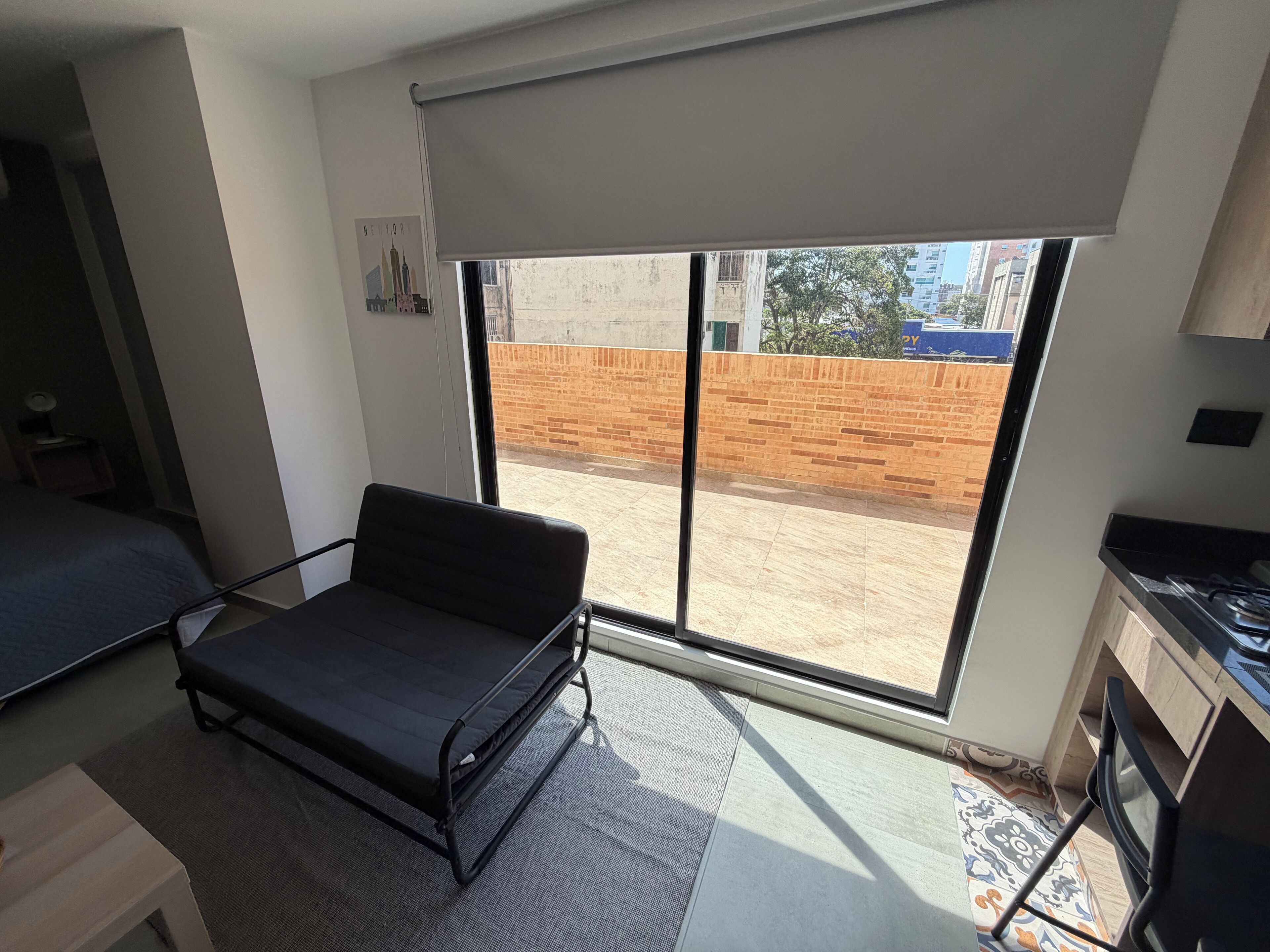 A compact room with a bed, a small table, a kitchenette, and a large window offering a view of a brick wall and buildings.