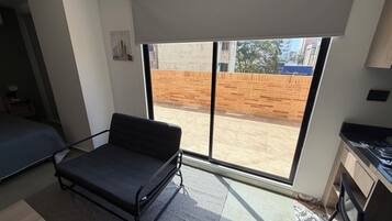 Habitación pequeña con cama, una mesita, kitchenette y un ventanal que ofrece vistas a un muro de ladrillo y edificios.