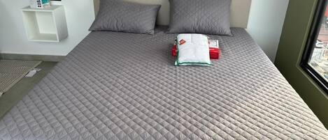 A neatly made bed with a quilted grey bedspread, two pillows, and a small bedside table with a box and a remote control.