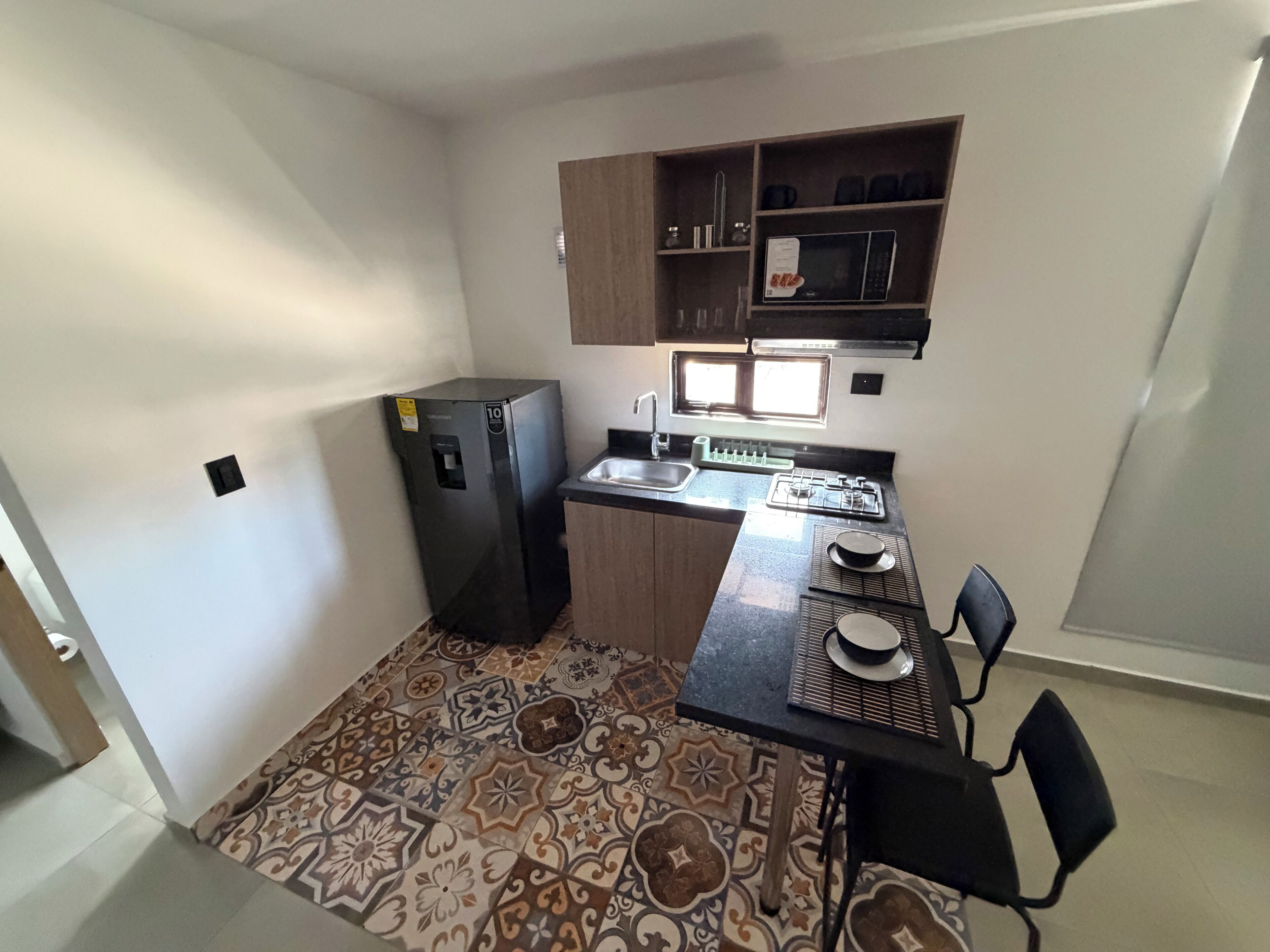 A compact kitchen with a refrigerator, sink, microwave, and a small dining area with a chair.