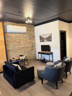 Ein modernes Wohnzimmer mit Backsteinwand, Flachbildfernseher, Couchtisch aus Holz und schwarzen sowie grauen Möbeln.