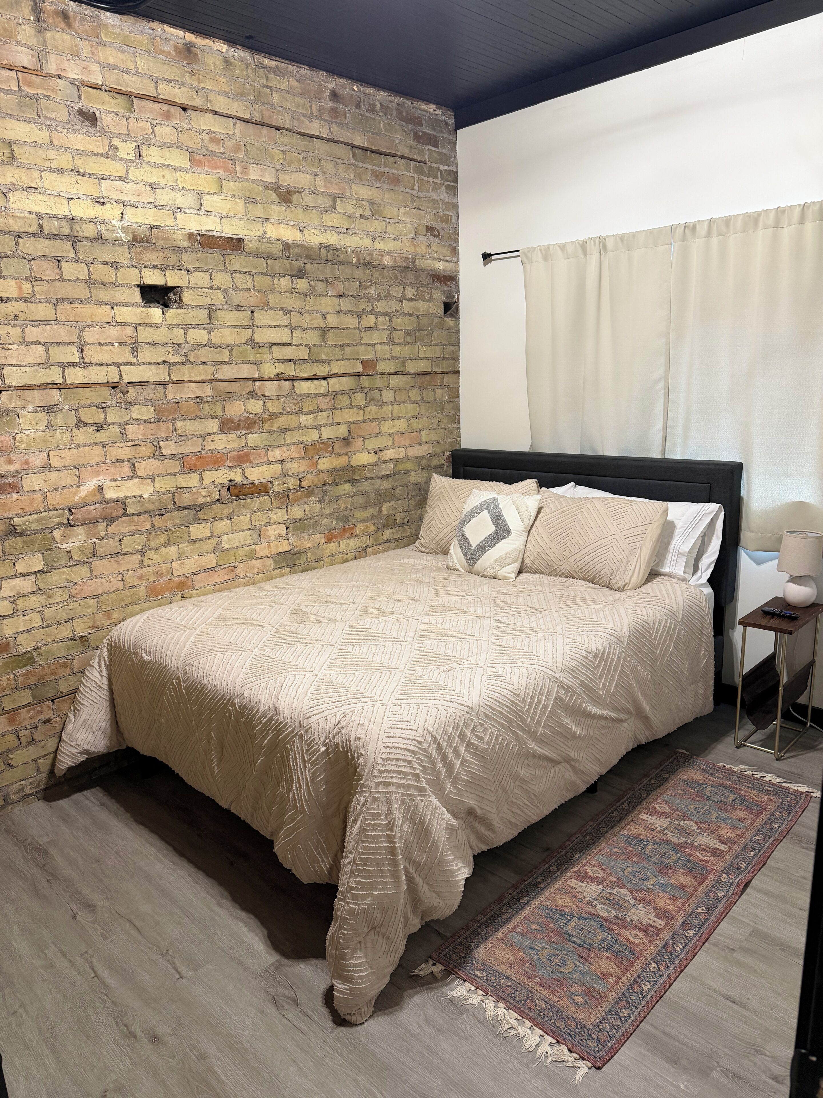 Una camera da letto con una parete in mattoni, un letto con copriletto decorato, un tappeto e un comodino con una lampada.