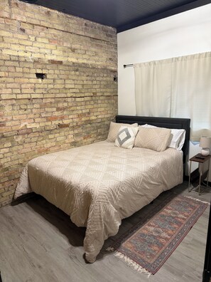A bedroom with a brick wall, a bed with a patterned bedspread, a rug, and a nightstand with a lamp.