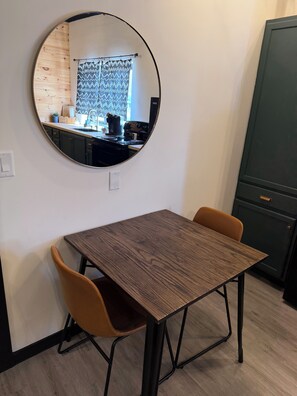 Een eetruimte met een ronde spiegel, een houten tafel, twee stoelen, en een keuken die door de spiegel zichtbaar is.