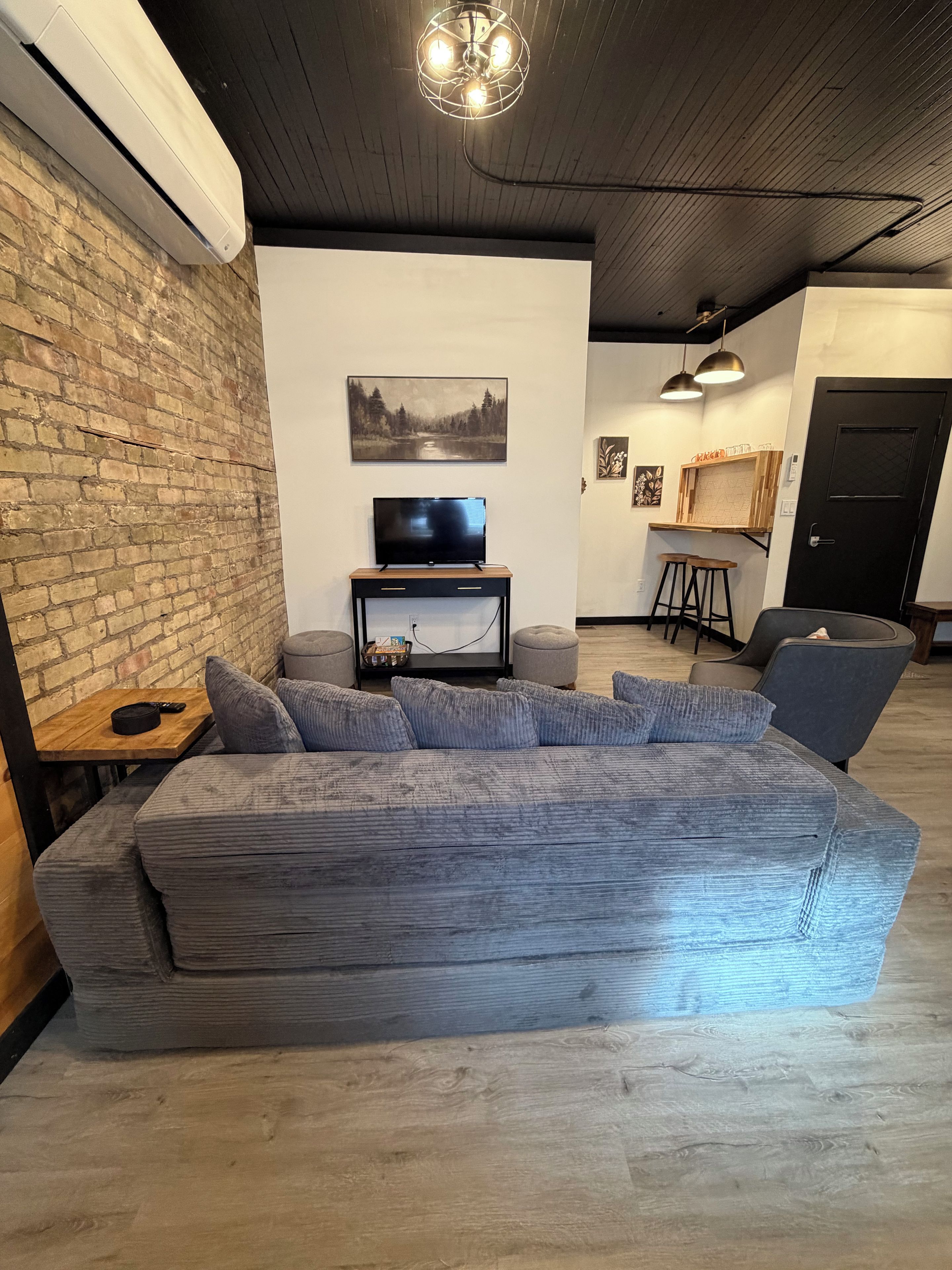 Un salón moderno con un amplio sofá seccional gris, un televisor de pantalla plana sobre un soporte de madera, una pared de ladrillo y un techo con tuberías a la vista.