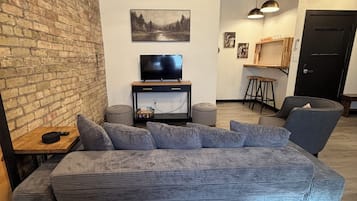 Un salón moderno con un amplio sofá seccional gris, un televisor de pantalla plana sobre un soporte de madera, una pared de ladrillo y un techo con tuberías a la vista.