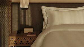 A neatly made bed with a pillow, a bedside table with a clock and a book, and a textured wall with a geometric pattern. - Rosewood AMAALA (Al Wajh)