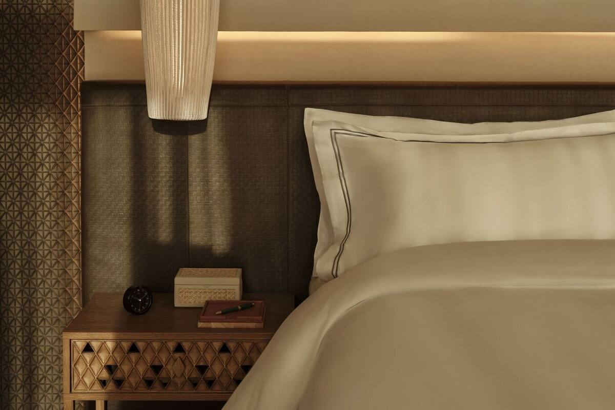 A neatly made bed with a pillow, a bedside table with a clock and a book, and a textured wall with a geometric pattern.
