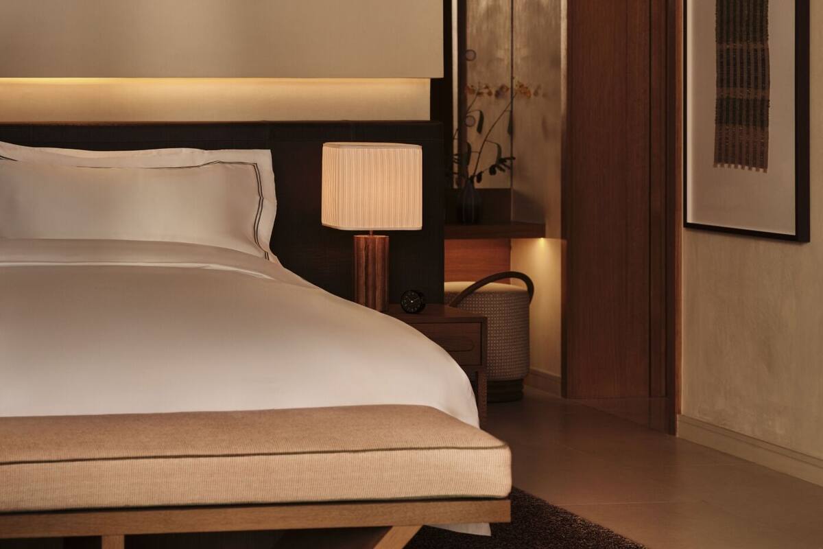 A neatly made bed with a headboard, a bedside lamp, a small table with a clock, and a framed picture on the wall.