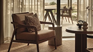 A chair with a cushion, a round table with a vase, and a view of the outdoors through large windows. - Rosewood AMAALA (Al Wajh)