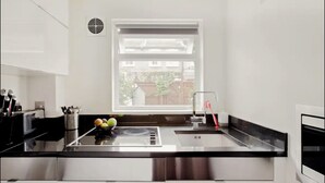Une cuisine moderne avec un plan de travail noir, des appareils en acier inoxydable, une fenêtre avec des stores et un panier de fruits sur l’évier.