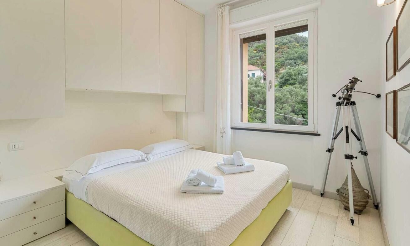 A bedroom with a bed, white curtains, a window with a view, a telescope, and a small table.