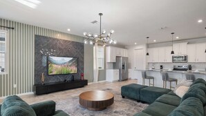 A modern living room with a large TV, green sofas, a round coffee table, and a kitchen area with white cabinets and a central island. - 11BR FamilyHome/Games @ Solara + Resort Access! (Kissimmee)