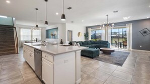 A modern kitchen with a central island, a dining area with a round table, and a living room with a sofa and a large window. - 11BR FamilyHome/Games @ Solara + Resort Access! (Kissimmee)