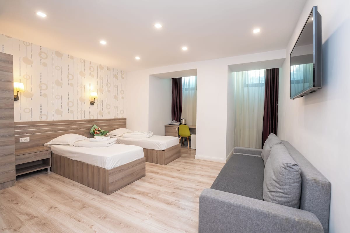 A modern hotel room with two beds, a sofa, a flat-screen TV, and a desk with a chair.