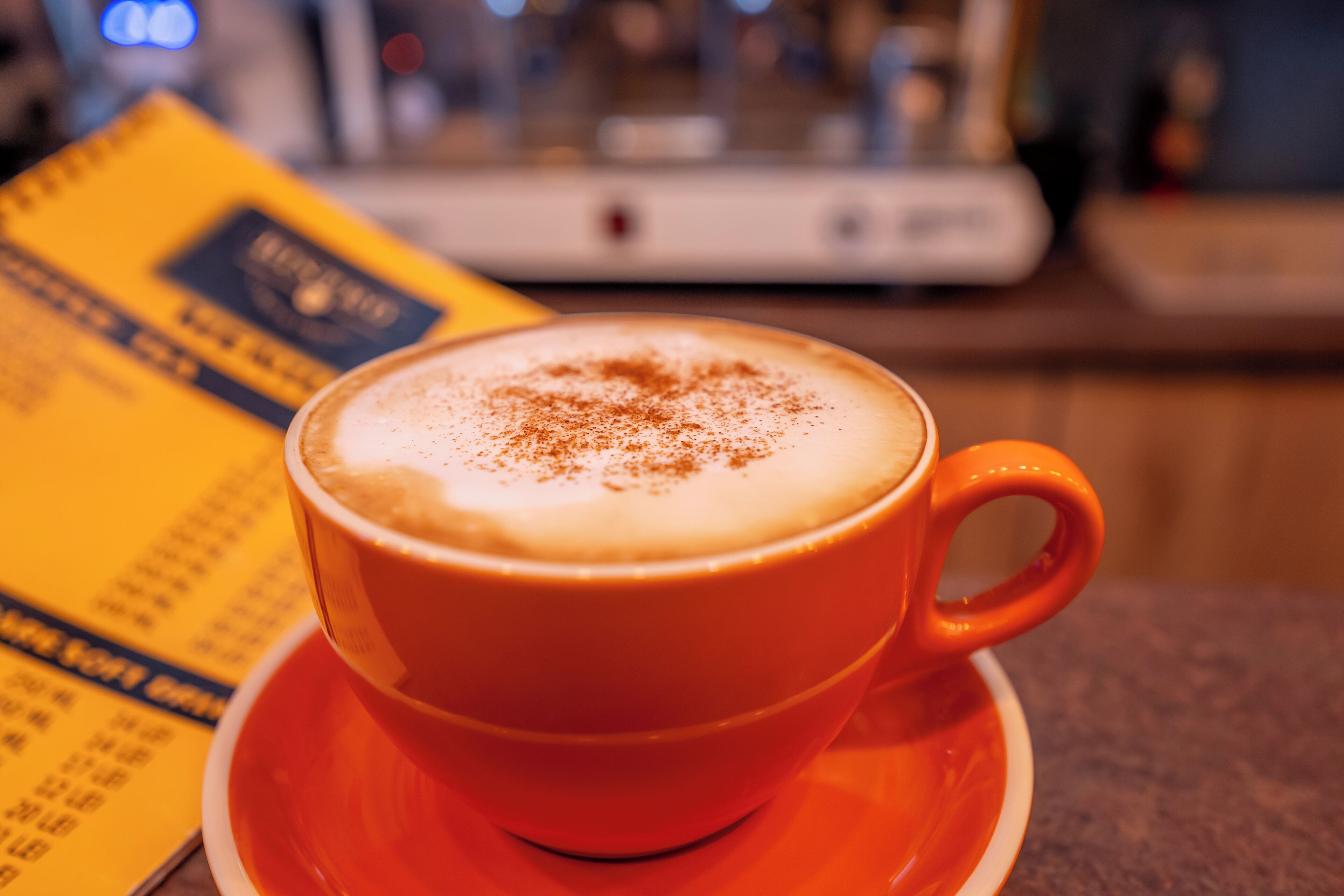 A cup of coffee with a frothy top on a saucer, placed on a table with a menu in the background.