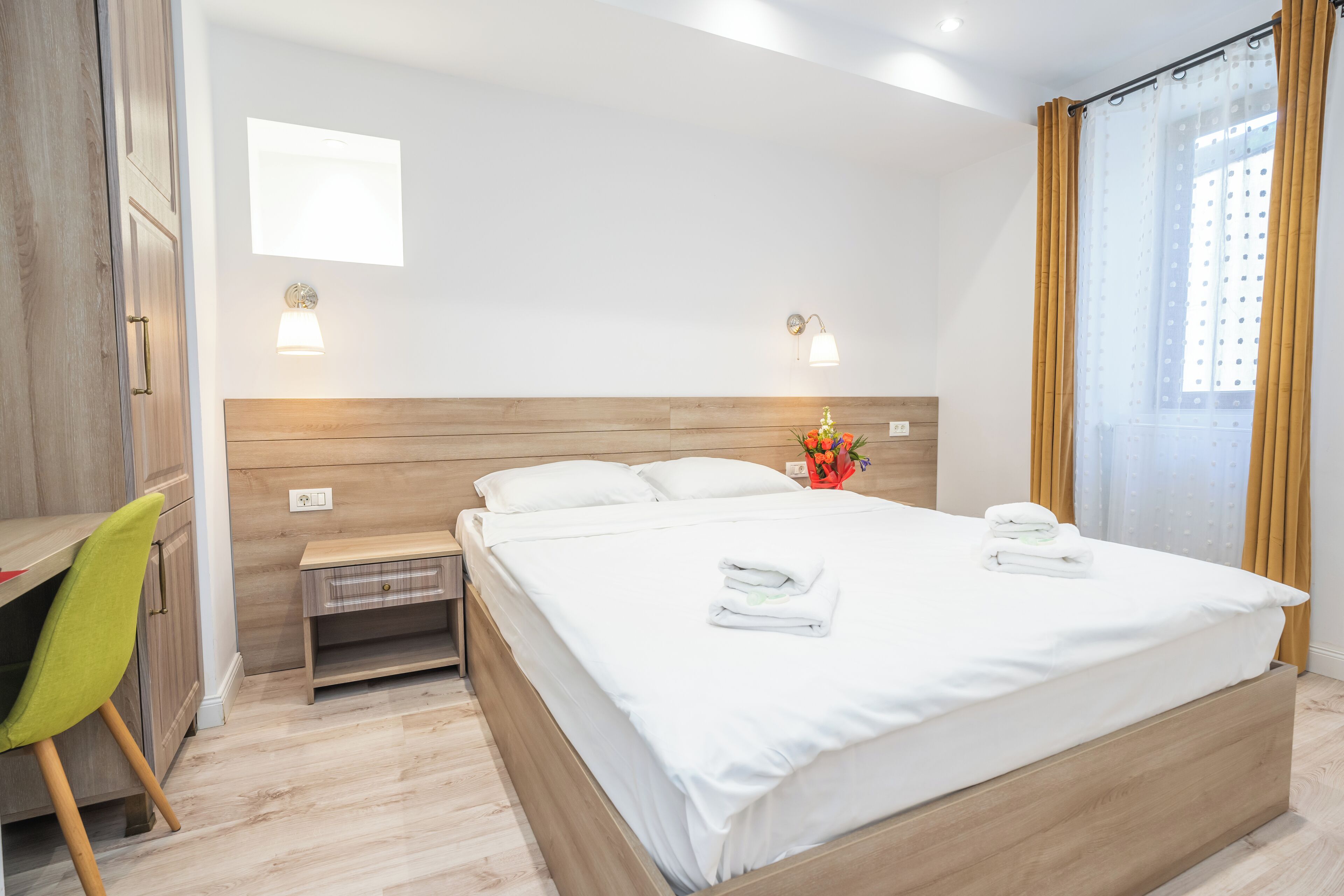 Quarto de hotel com cama de madeira, uma escrivaninha com cadeira, duas luminárias de cabeceira e uma janela com cortinas.