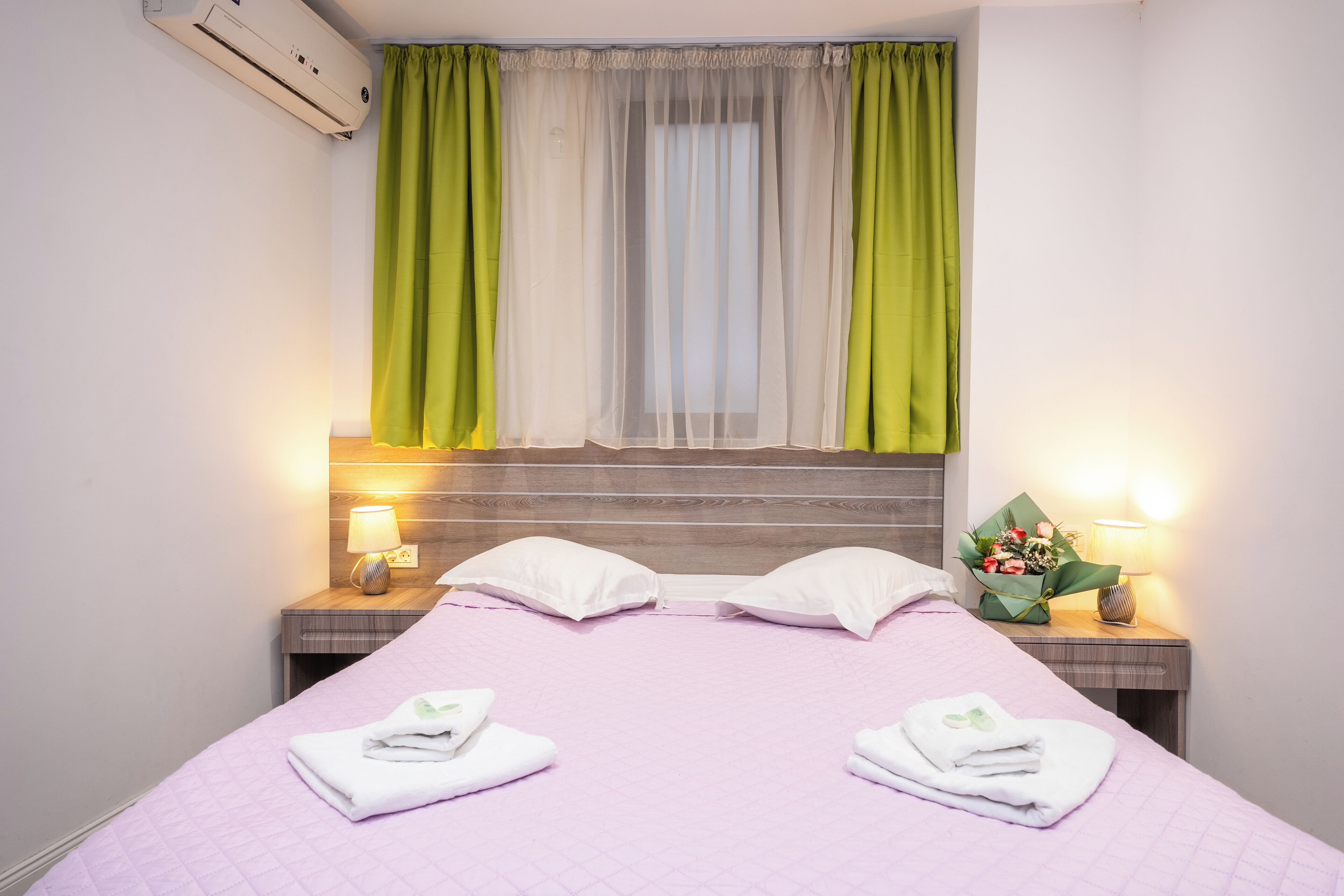 Una habitación de hotel con una cama, dos mesitas de noche, un ventanal con cortinas translúcidas y un aire acondicionado instalado en la pared.