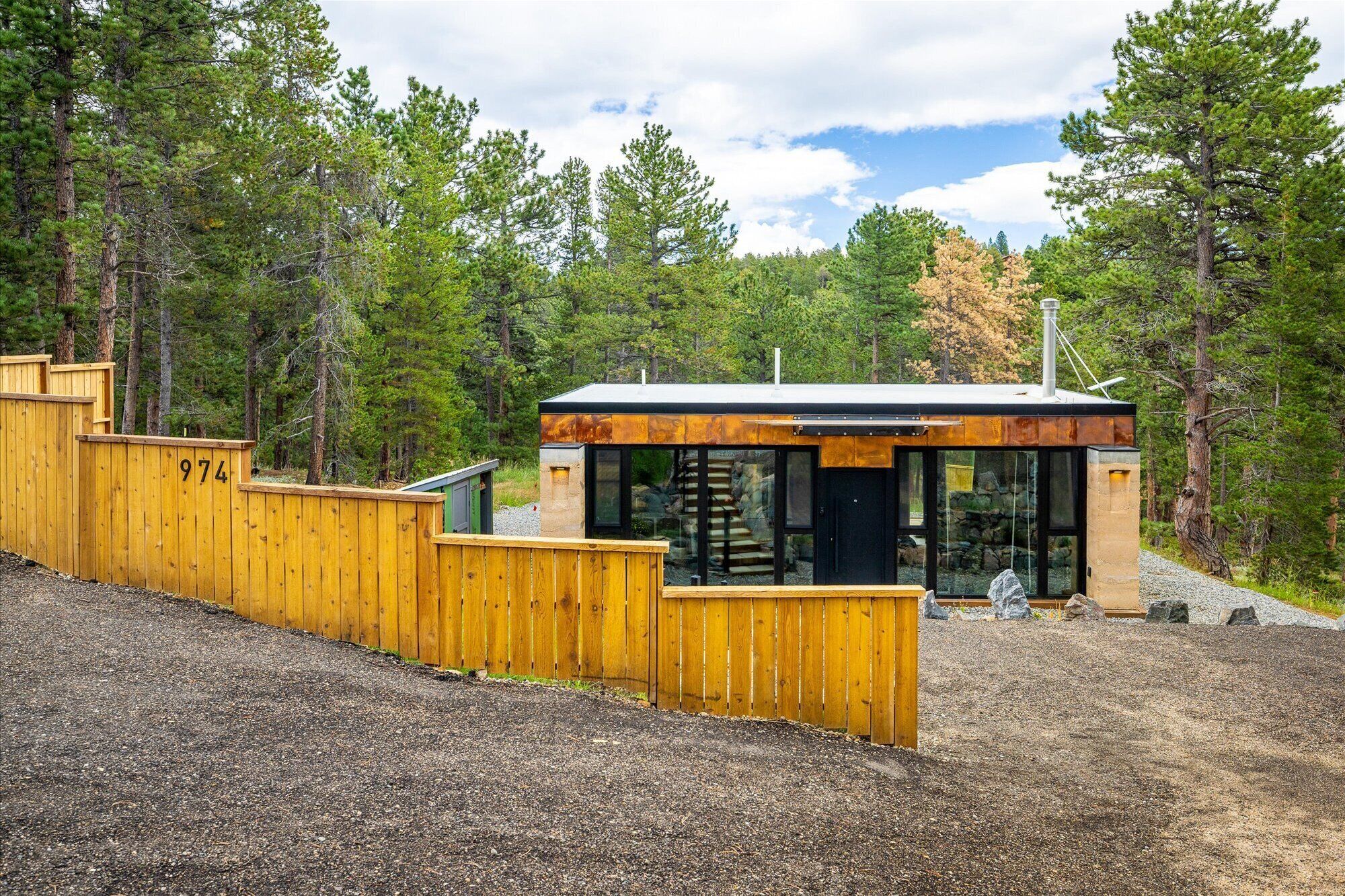 Une cabane moderne, dotée d’une clôture en bois et portant le numéro « 974 », est entourée d’une forêt.