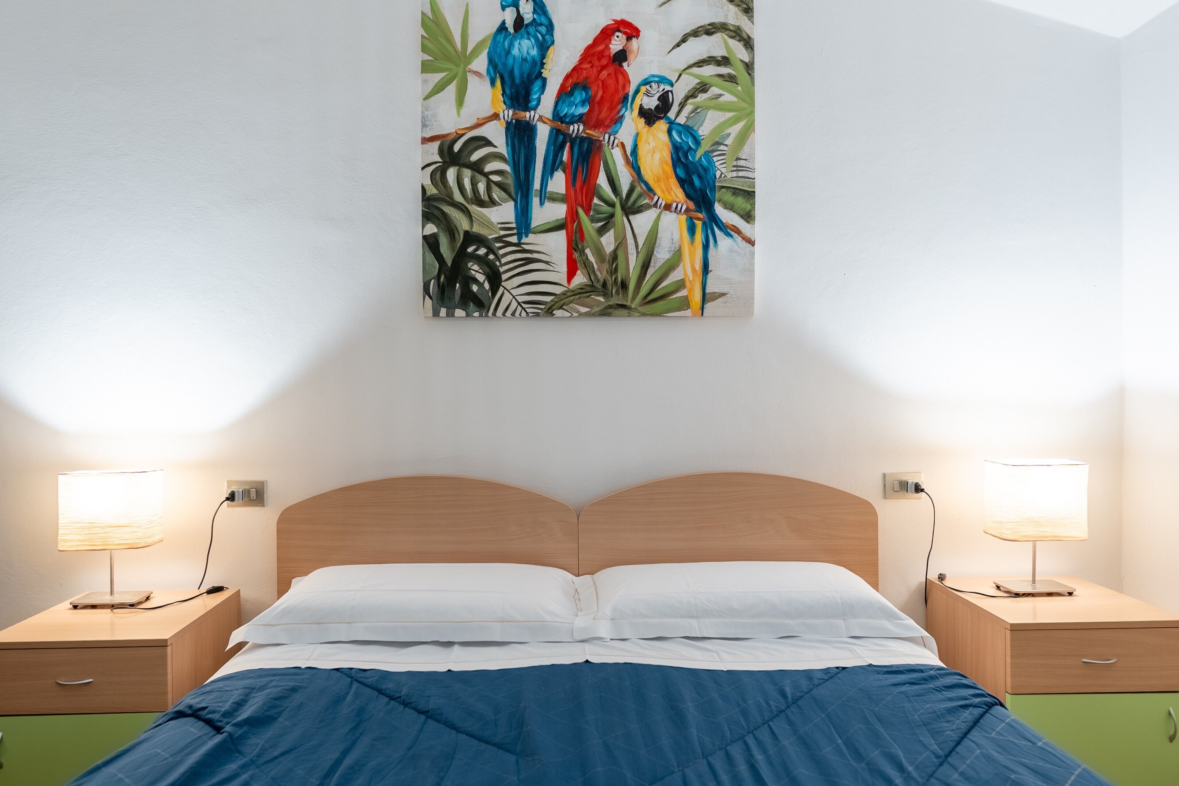 Une chambre à coucher avec une tête de lit en bois, un lit recouvert d’une couette blanche et bleue, deux tables de chevet avec des lampes, et un tableau représentant des perroquets colorés accroché au mur.