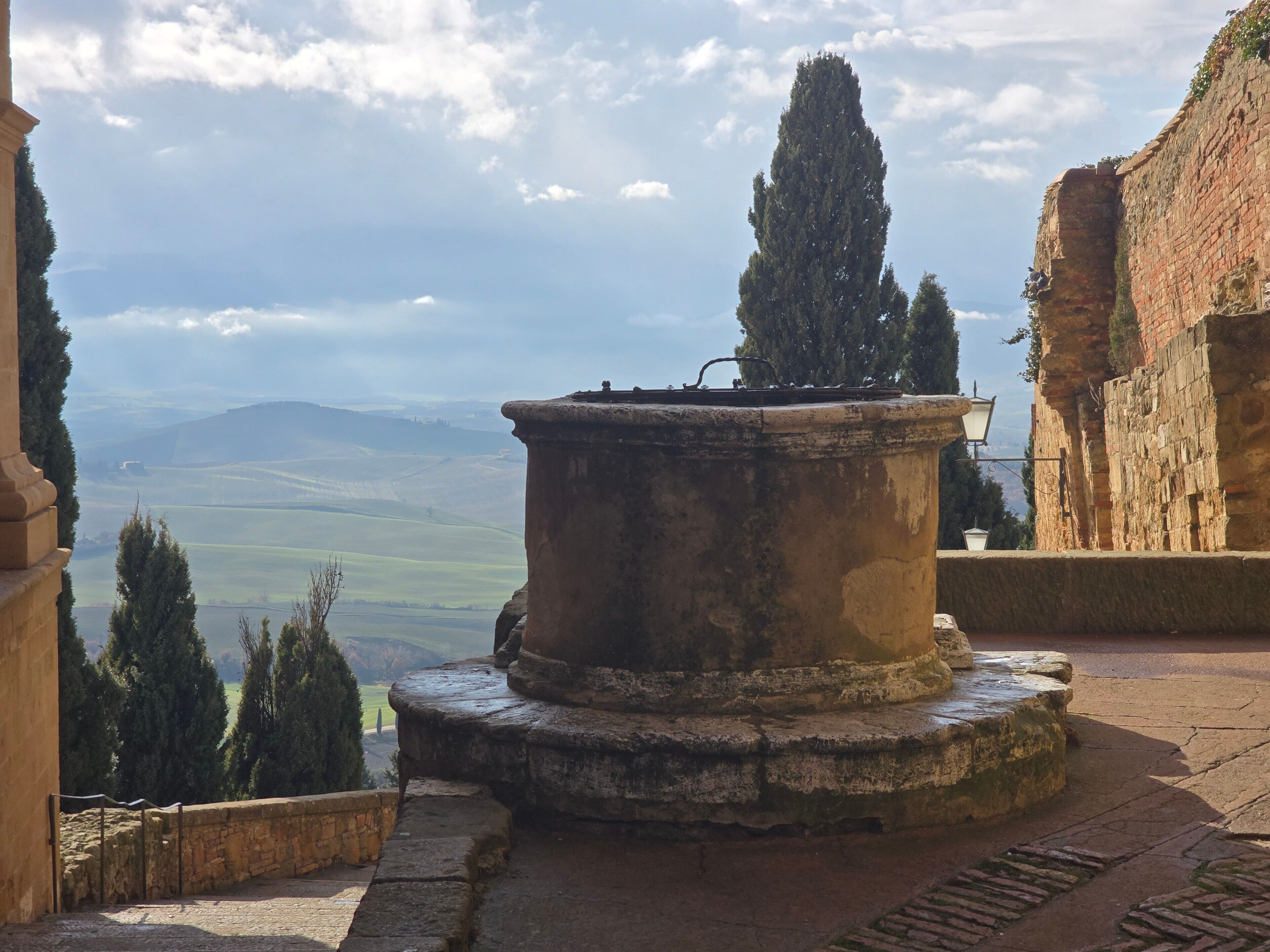 An outdoor courtyard with a stone fountain, a tall tree, and a view of distant hills.