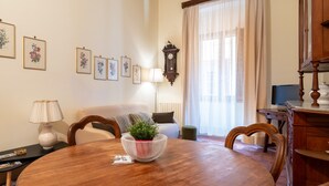 Un comedor con una mesa redonda de madera, sillas, un florero con planta, un cuadro de flores en la pared, una lámpara, un sofá, un reloj y un televisor.