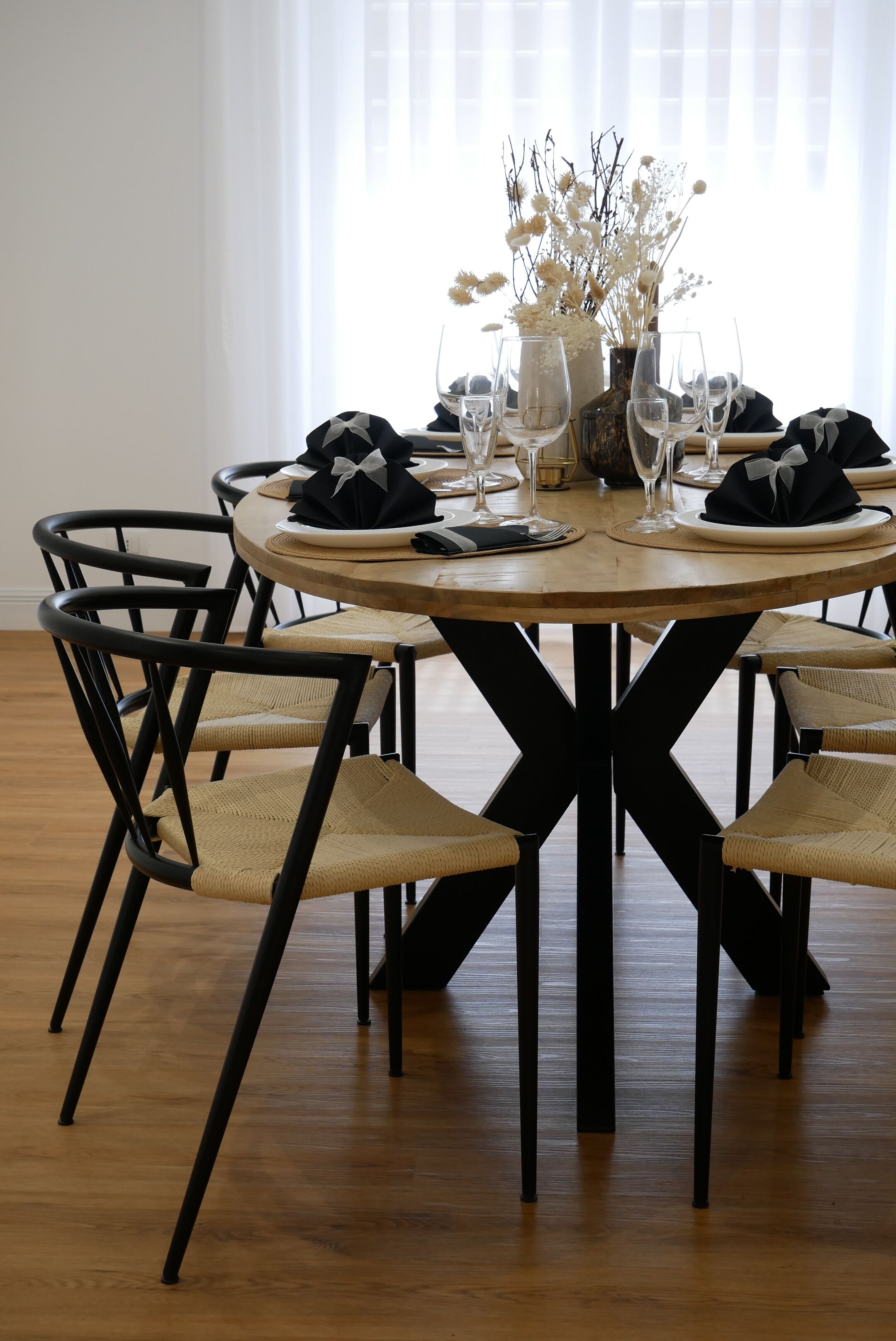 Une table de salle à manger ronde, entourée de chaises noires, un vase contenant des fleurs séchées et des verres à vin disposés pour six personnes.