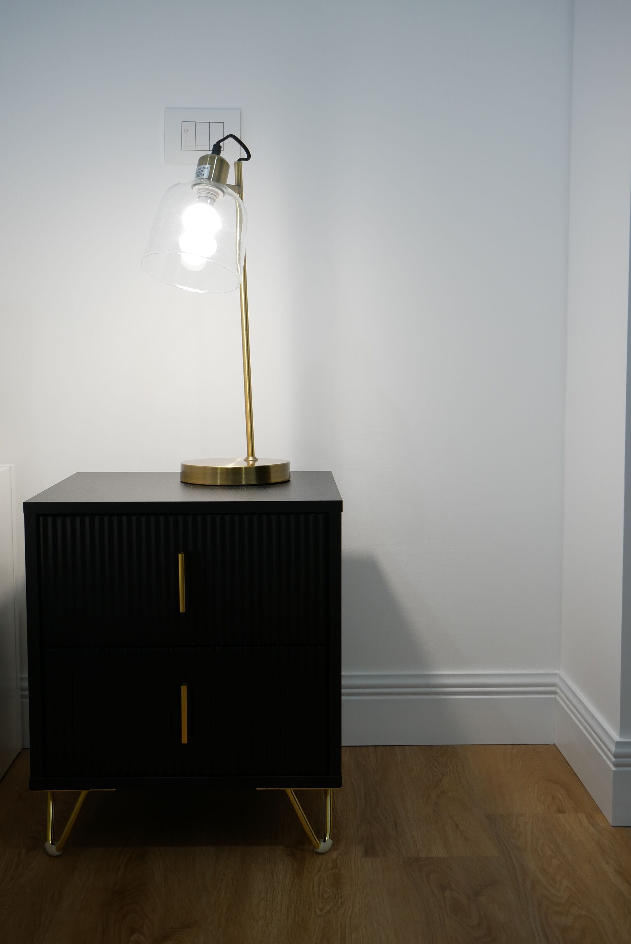 A black nightstand with a clear glass lamp and a gold base, positioned against a white wall with a light switch above it.