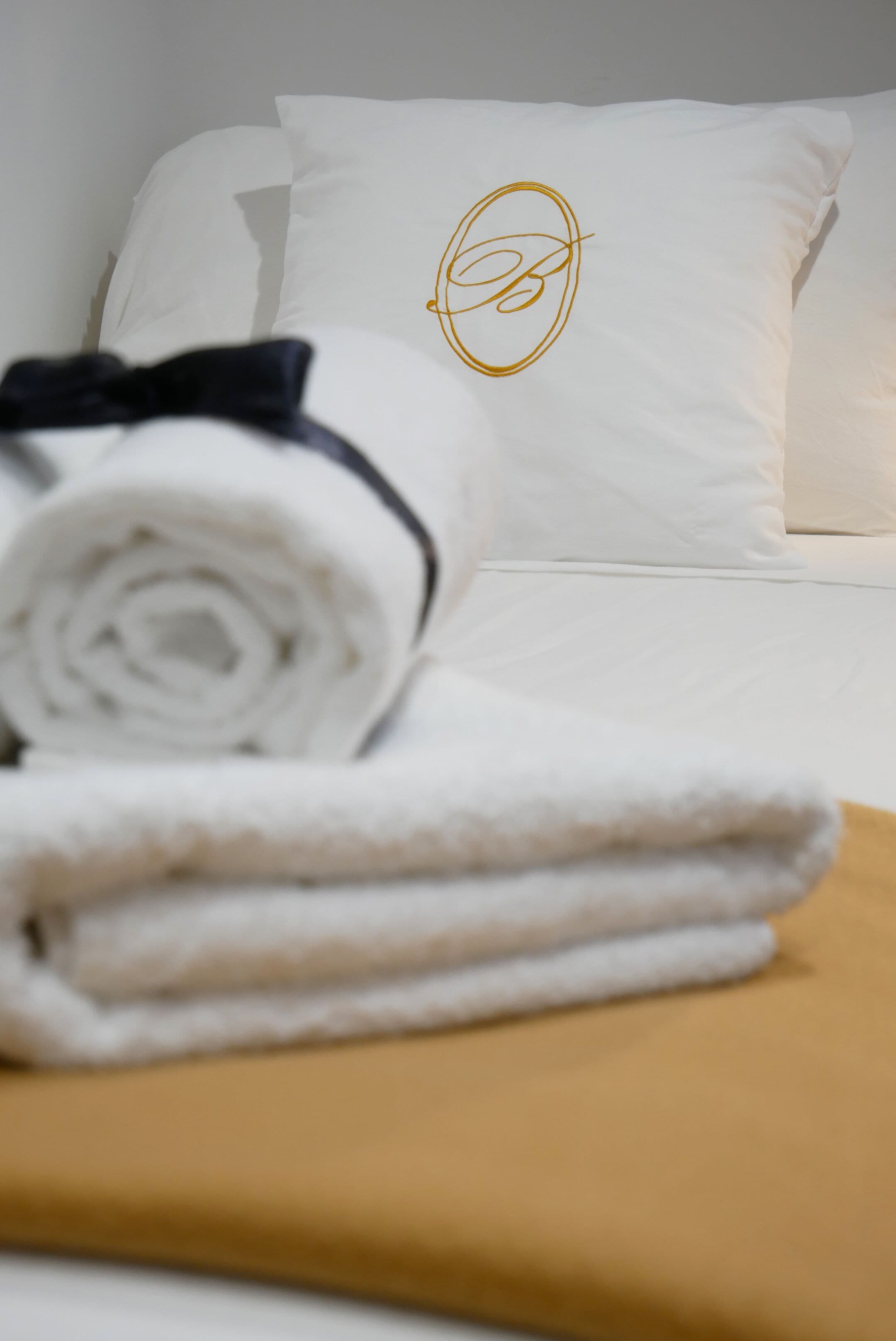 A neatly made bed with white linens and a black bow, a white pillow with a gold monogram, and a folded towel on a mustard-colored blanket.