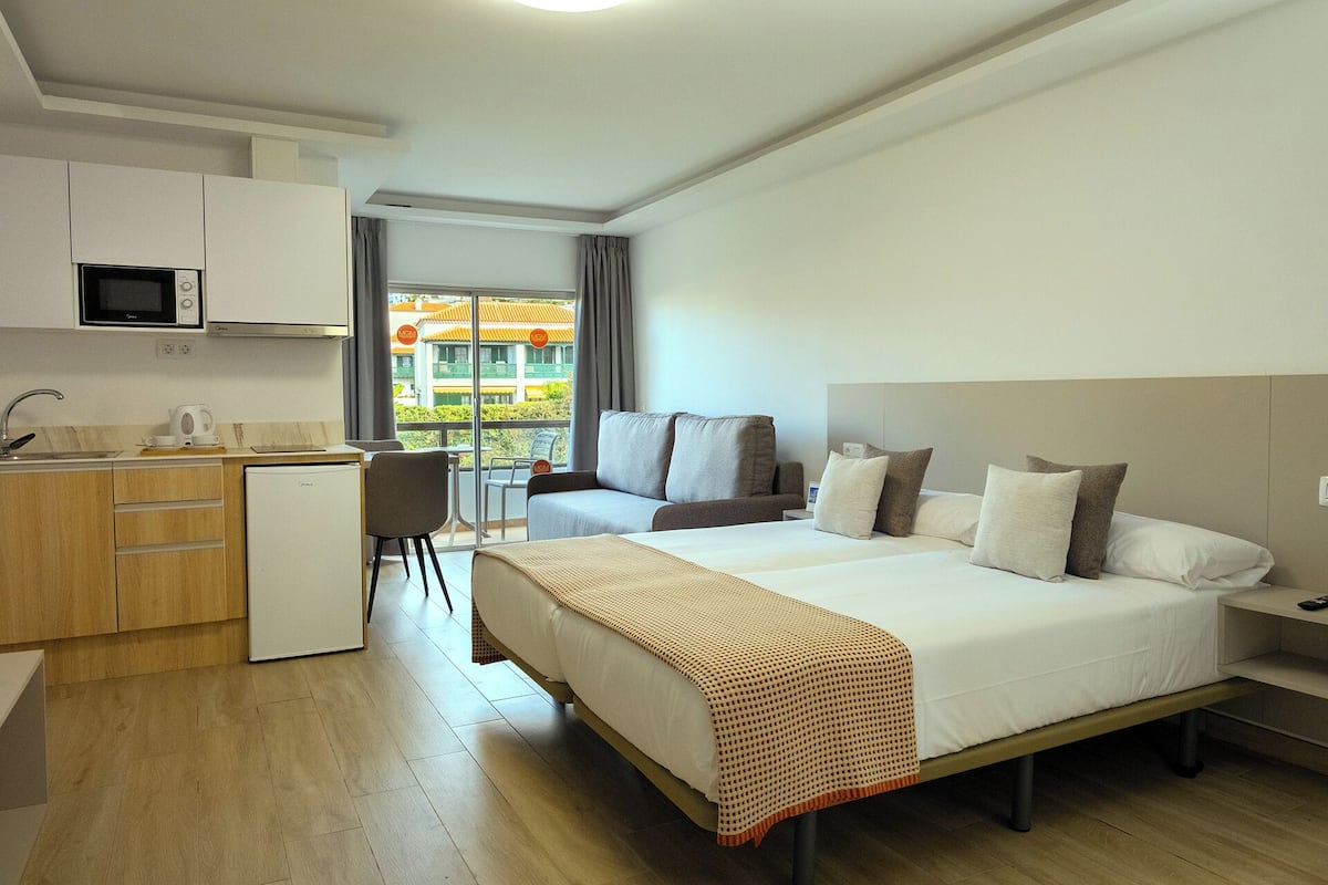Ein Hotelzimmer mit Bett, Schreibtisch, Stuhl, einer Küchenzeile und Blick auf umliegende Gebäude.
