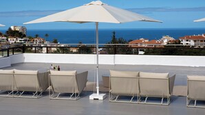 Een terras op het dak met een grote witte parasol, beige lounge stoelen en uitzicht op zee en gebouwen.