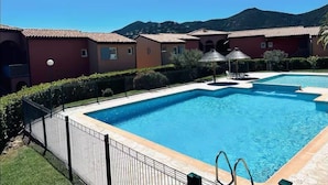 Una piscina con solárium empedrado, rodeada por una valla metálica, junto a un edificio con fachada roja y el paisaje de una montaña al fondo.
