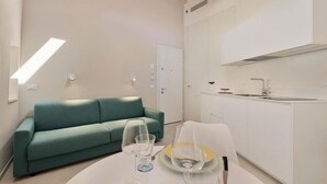 Ein modernes, minimalistisches Wohnzimmer mit einem türkisfarbenen Sofa, einem Esstisch mit Weingläsern und Tellern sowie einer Küchenzeile mit weißen Schränken und einer Spüle.