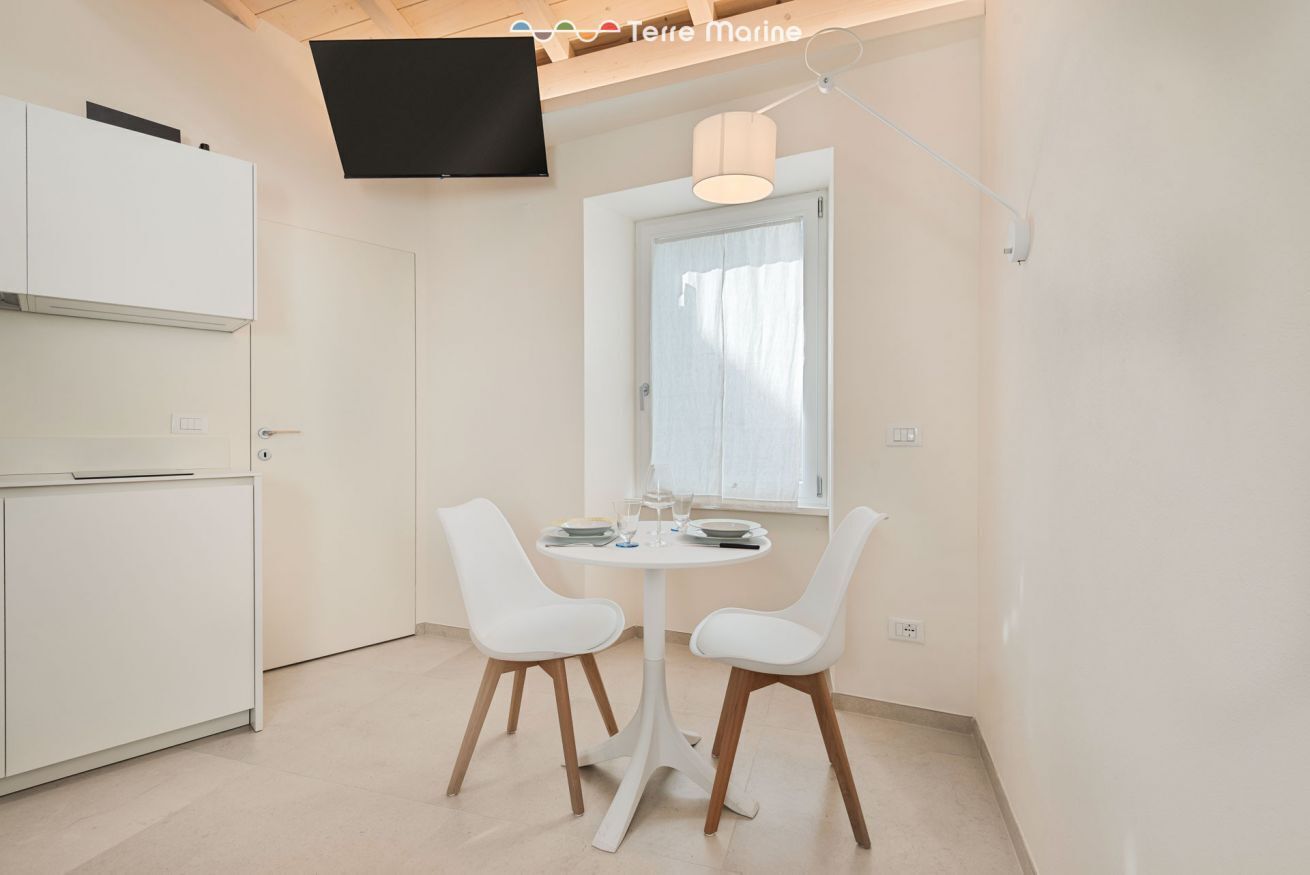 A modern kitchen with white cabinetry, a flat-screen TV mounted above the counter, and a dining area with two white chairs and a small round table.