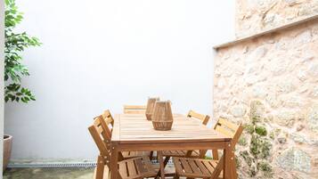 A wooden table with six chairs and a vase in the center, set against a white wall and a stone wall.