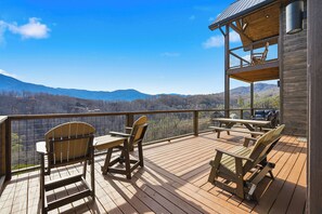Eine Holzterrasse mit Gartenmöbeln, Blick auf Berge und ein Gebäude mit Balkon.