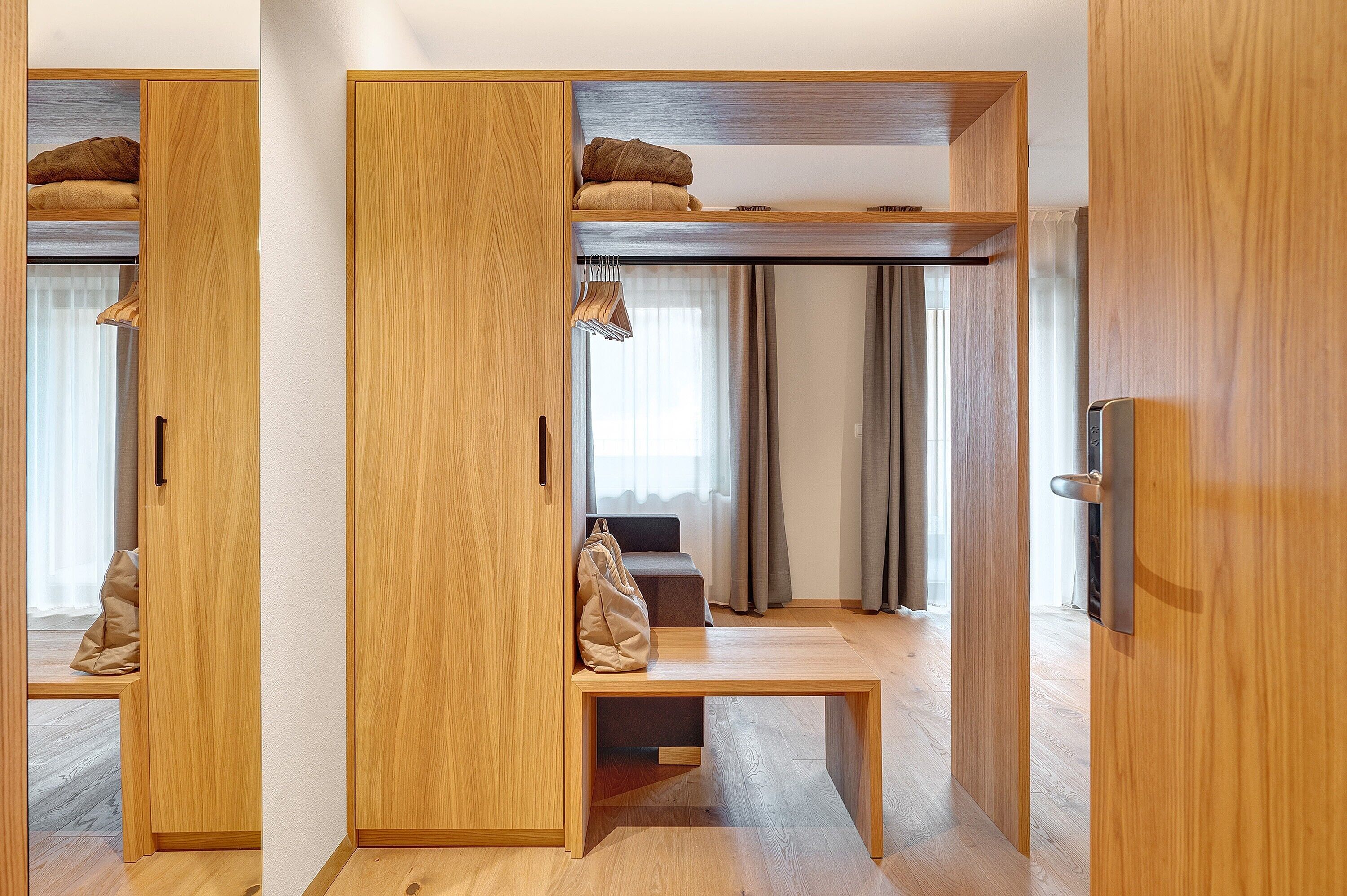 Ein Zimmer mit Holzschrank, einer Sitzbank, einem Bett mit Kopfteil und einem Fenster mit Vorhängen.