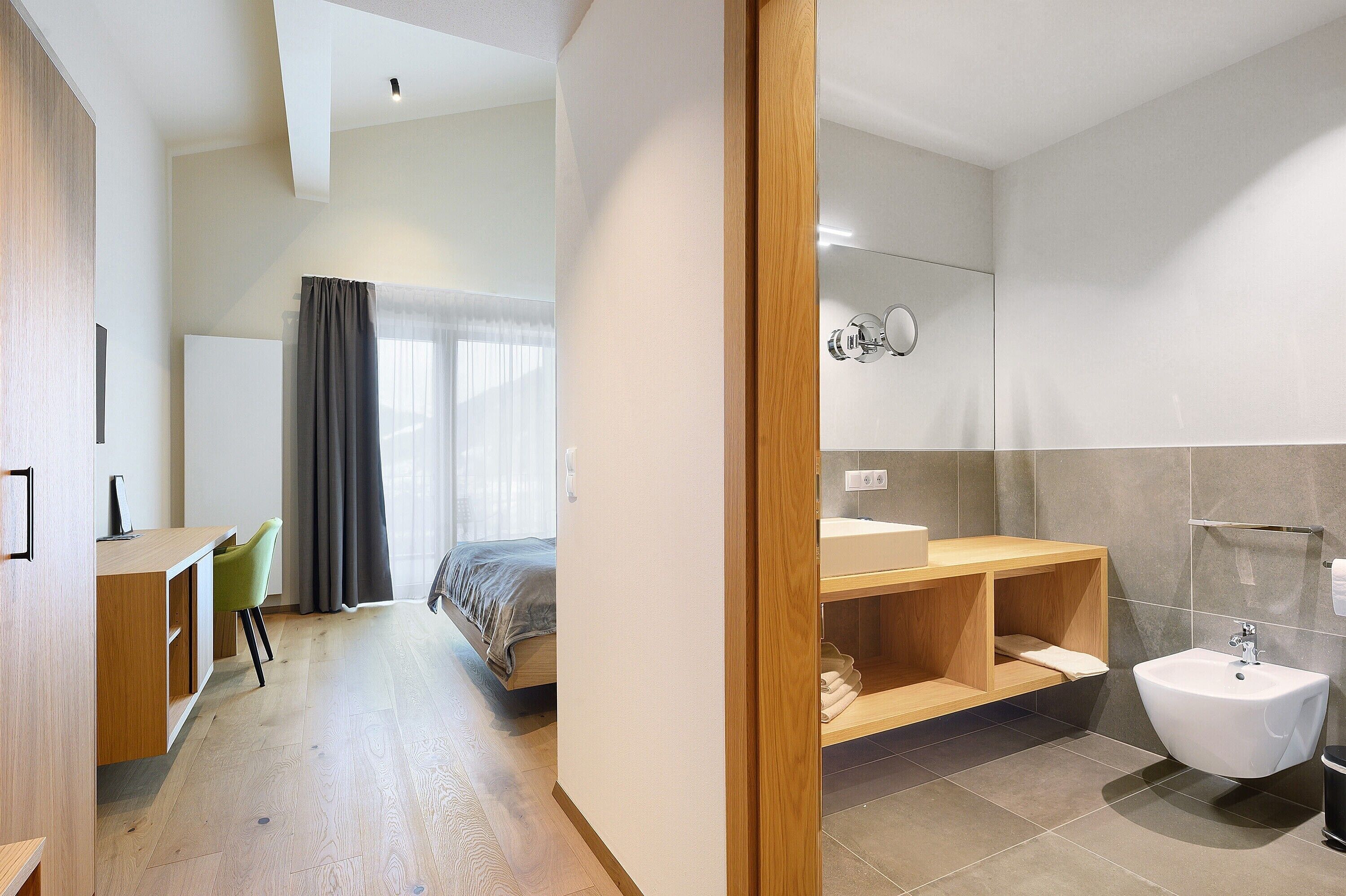 Ein modernes Hotelzimmer mit Schreibtisch aus Holz, einem Bett, einem Badezimmer mit Waschbecken und Bidet sowie einem großen Spiegel.