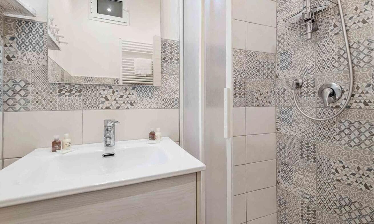 Un bany amb un lavabo blanc, un mirall i una dutxa amb un mur de rajoles estampades.