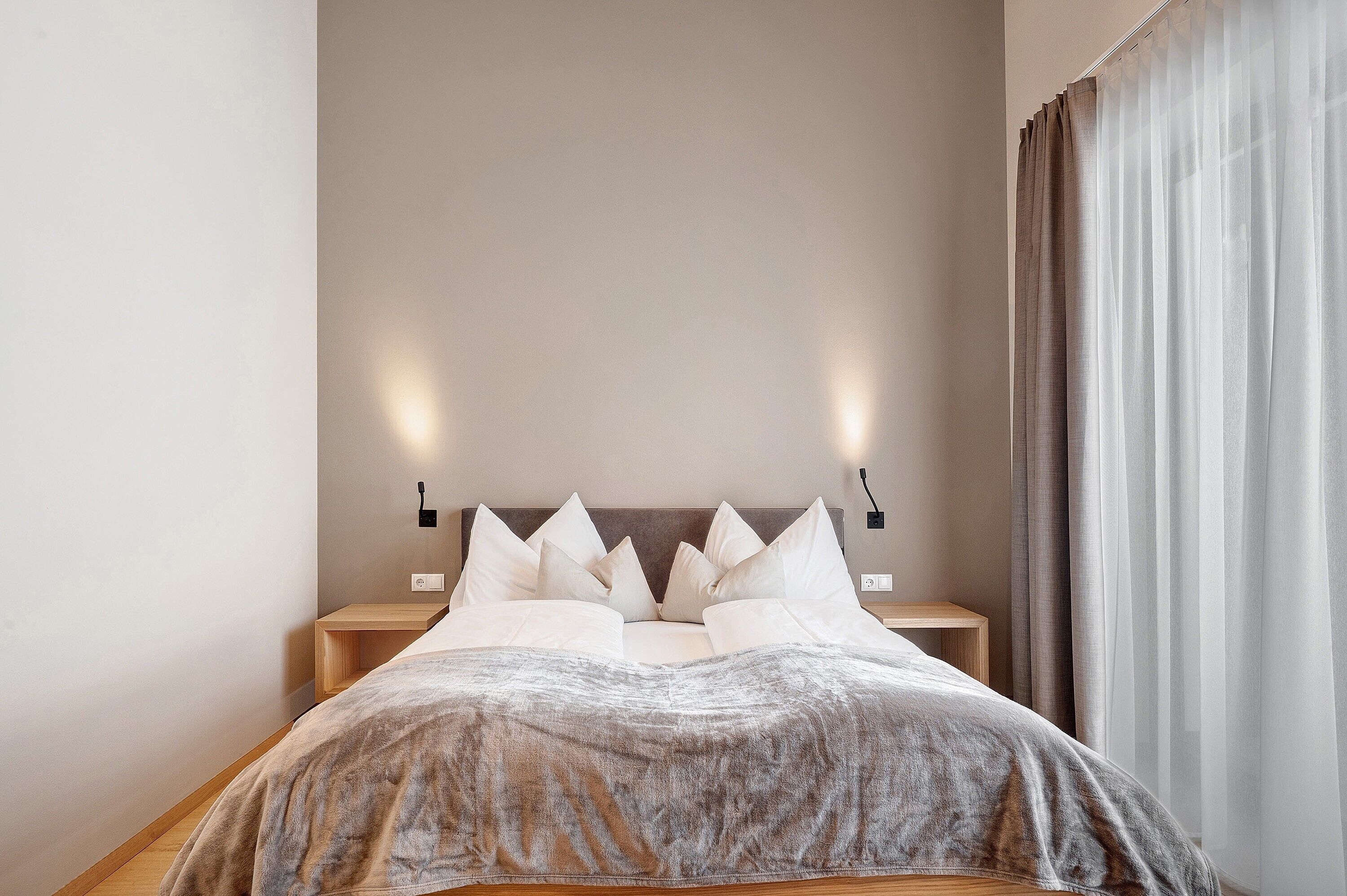 Un letto rifatto con un copriletto grigio, due comodini con lampade e una finestra con tende leggere.
