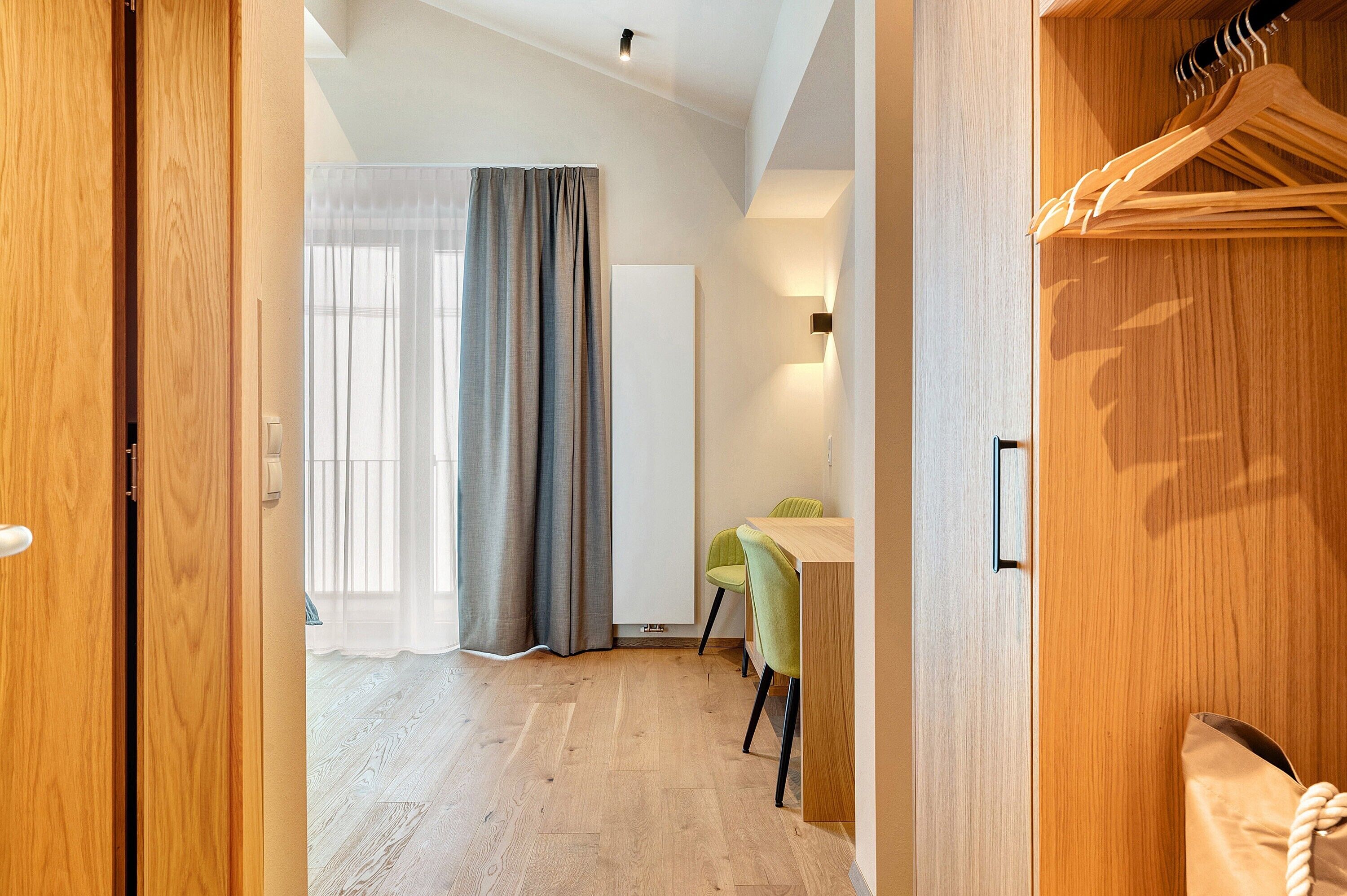 A hotel room with a wooden wardrobe, a desk with two chairs, a large window with curtains, and a light fixture on the wall.