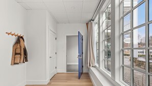 Un pasillo con suelo de madera, un perchero con abrigos, una puerta azul y ventanas amplias con vistas al exterior.