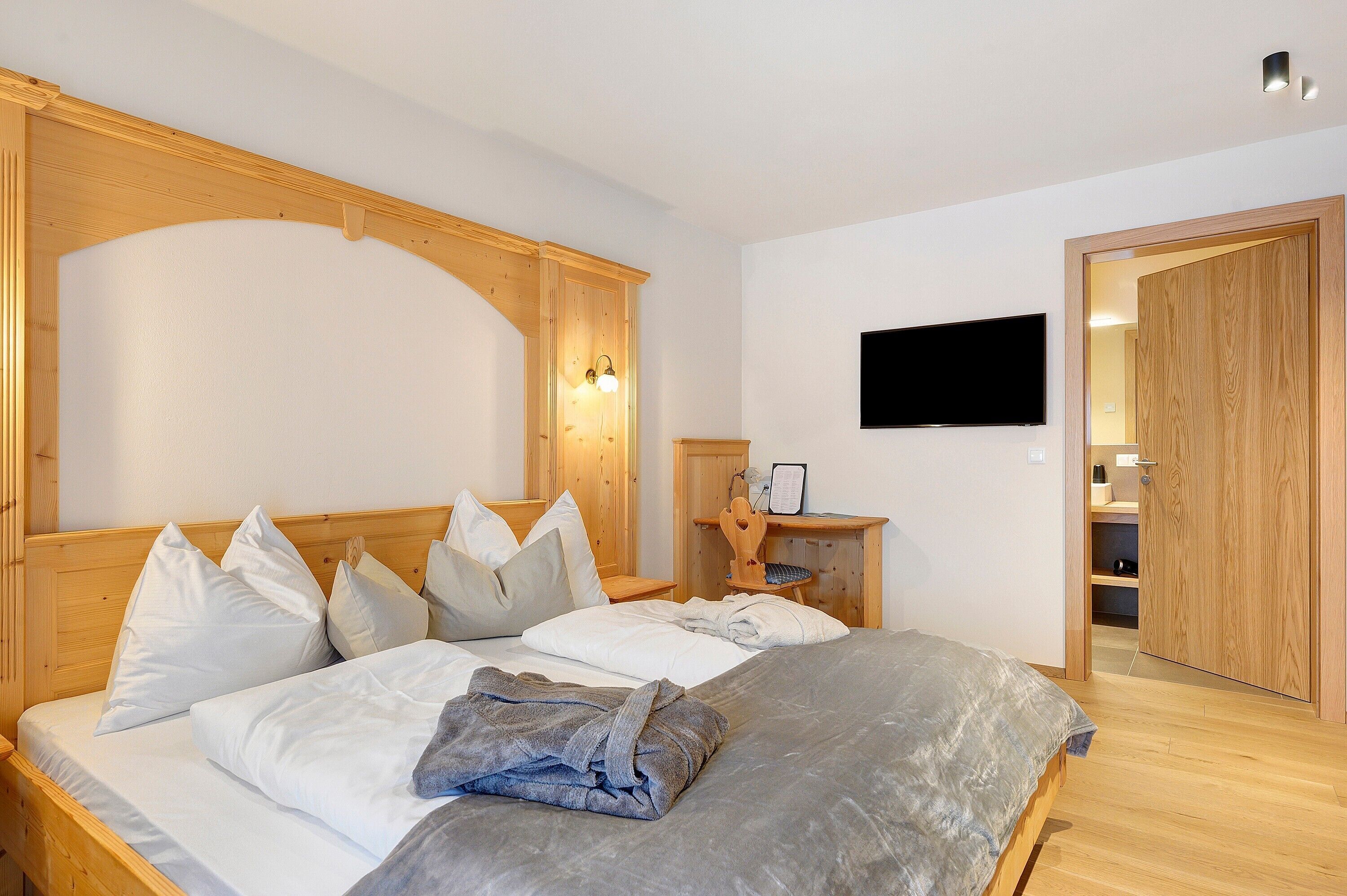 Un dormitorio con cabecera de madera, un televisor de pantalla plana montado en la pared, una mesita de noche con lámpara y un armario con espejo.